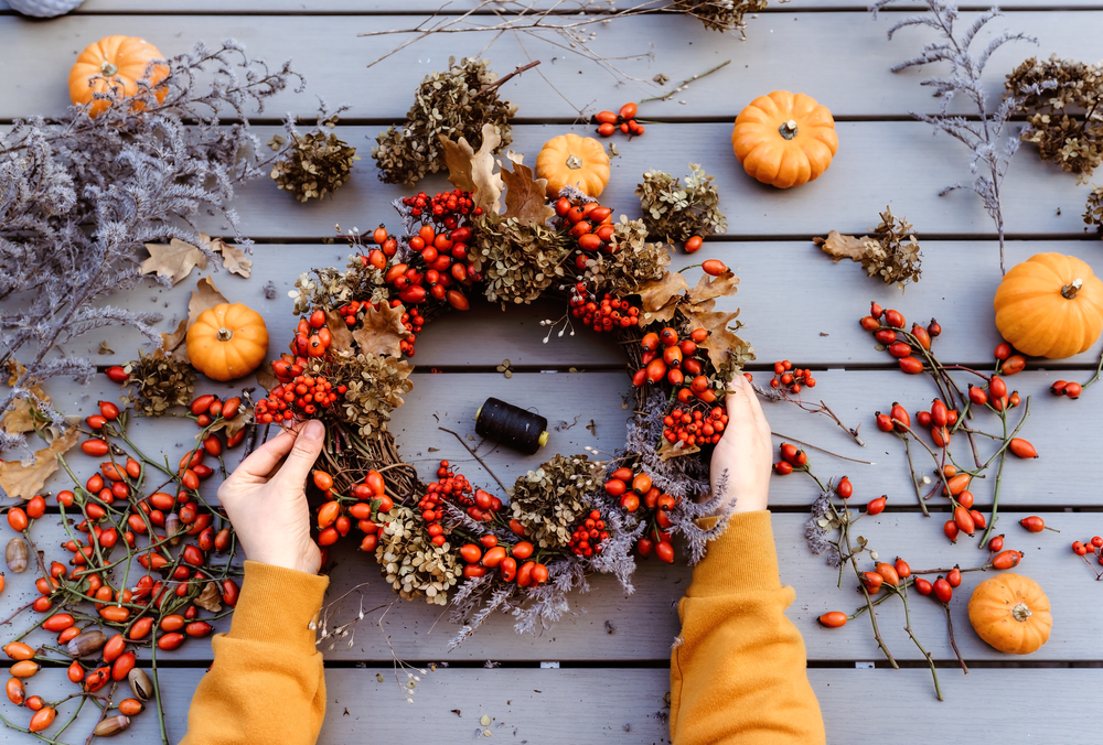 Girl making floral autumn door wreath using colorful rosehip berries, rowan, dry flowers and pumpkins. Fall flower decoration workshop, florist at work.